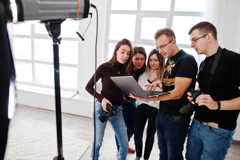 Photographer explaining about the shot to his team in the studio and looking on laptop. Talking to his assistants holding a camera during a photo shoot. Teamwork and brainstorm.